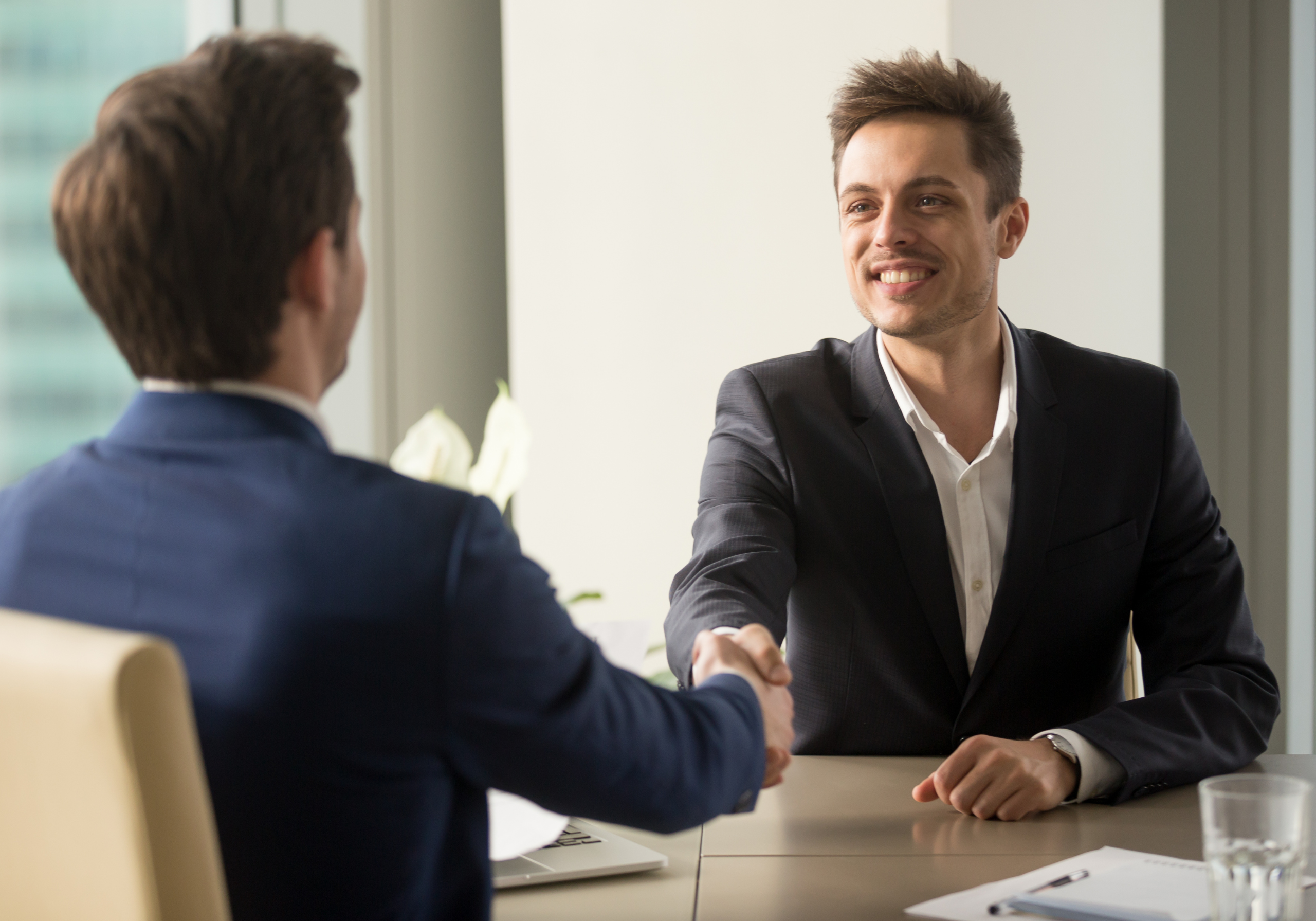 Due giovani uomini in abito formale si stringono la mano con un sorriso durante un colloquio di lavoro in un ufficio moderno e luminoso.